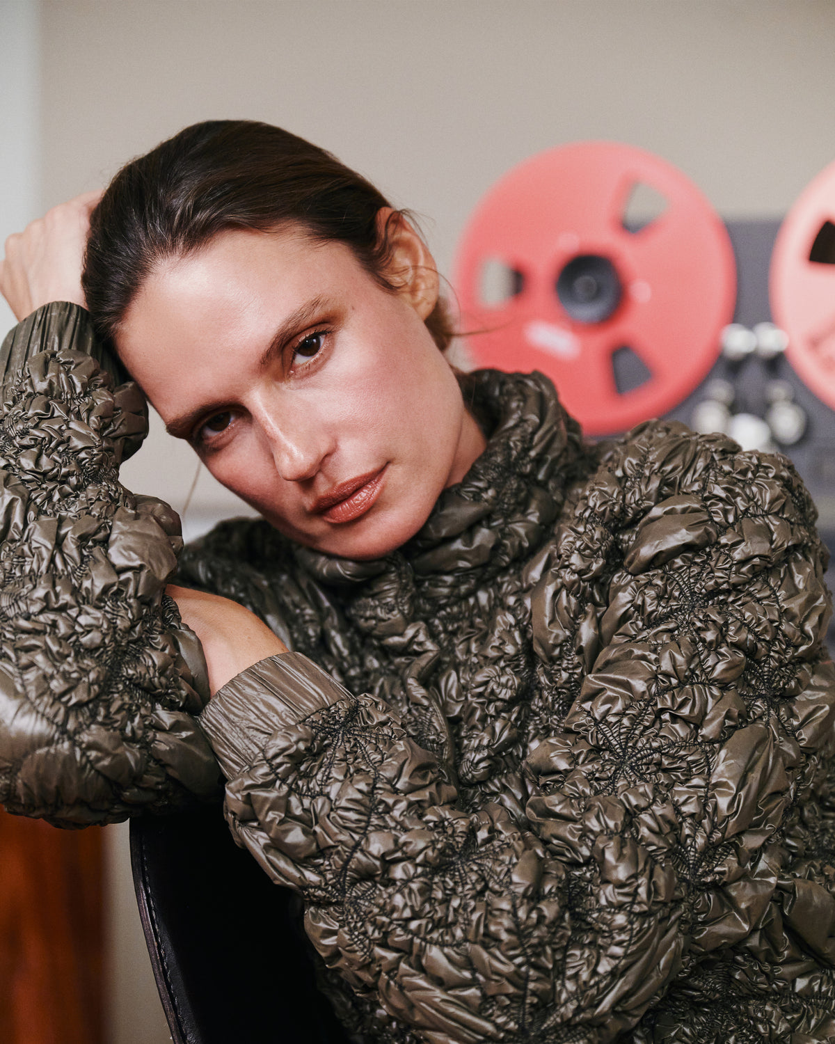 A close-up portrait of a woman wearing a heavily textured, crinkled dark brown or bronze turtleneck sweater, looking directly at the camera. The background is a soft-focus indoor setting with warm neutral tones and red circular decor.
