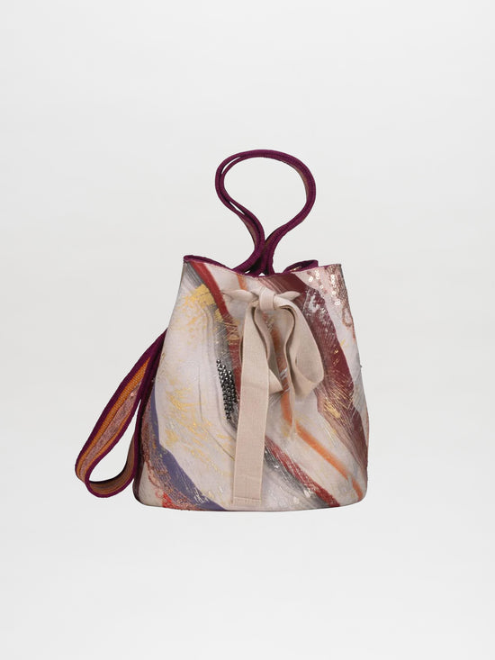 The Mochila Laaput Gradient Trinitaria is a multicolored, abstract-patterned tote bag featuring a purple handle and beige bow on the front, exemplifying exquisite Wayuu craftsmanship. With its unique design set against a plain white background, this mochila handbag effortlessly catches the eye.