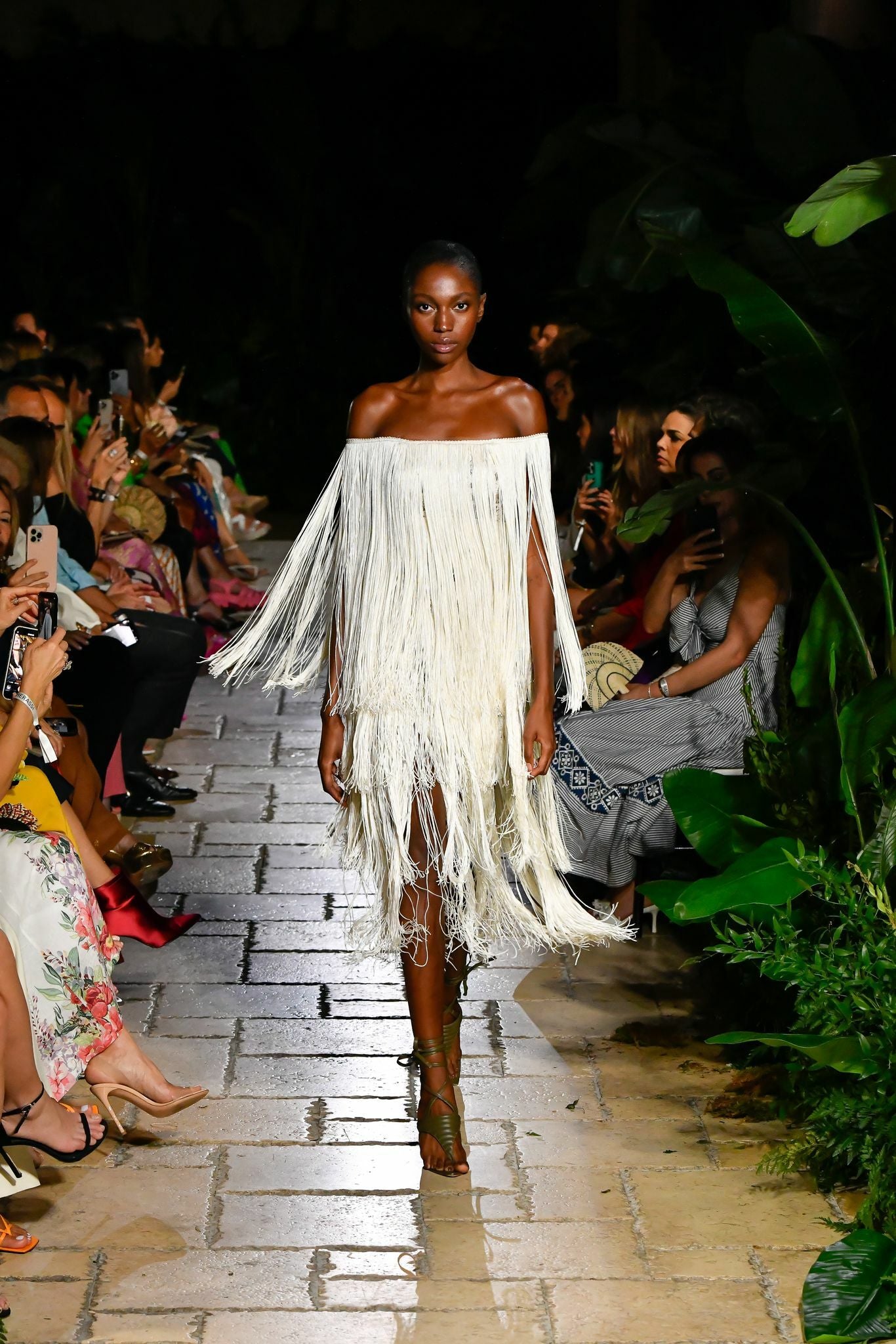 Model walking on a runway in a fringe off-shoulder dress, surrounded by seated audience capturing the moment with phones.