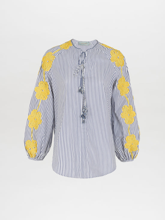 The Molveno Blouse Yellow Embroidered Flowers is a blue and white striped blouse adorned with yellow floral embroidery on the sleeves and shoulders, featuring a tied neckline and long, gathered sleeves. This piece exemplifies high fashion with its intricate details and elegant design.