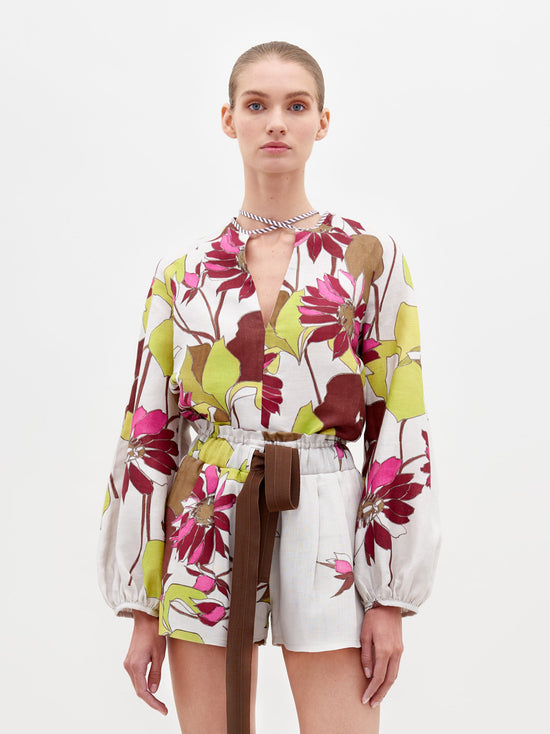 A person stands against a plain background, wearing the Molveno Blouse in Botanical Multifloral, featuring a floral-patterned long-sleeve top with a deep V-neckline and matching high-waisted shorts, cinched at the waist with a brown belt. Pre-order now to ensure it arrives by the February 15th, 2025 ship-by date.