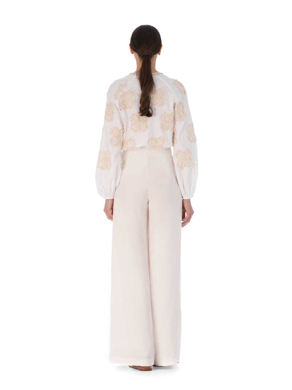 Rear view of a person with long hair wearing the Molveno Blouse Cream Flowers, featuring embroidered floral motifs, paired with high-waisted, wide-leg pants.