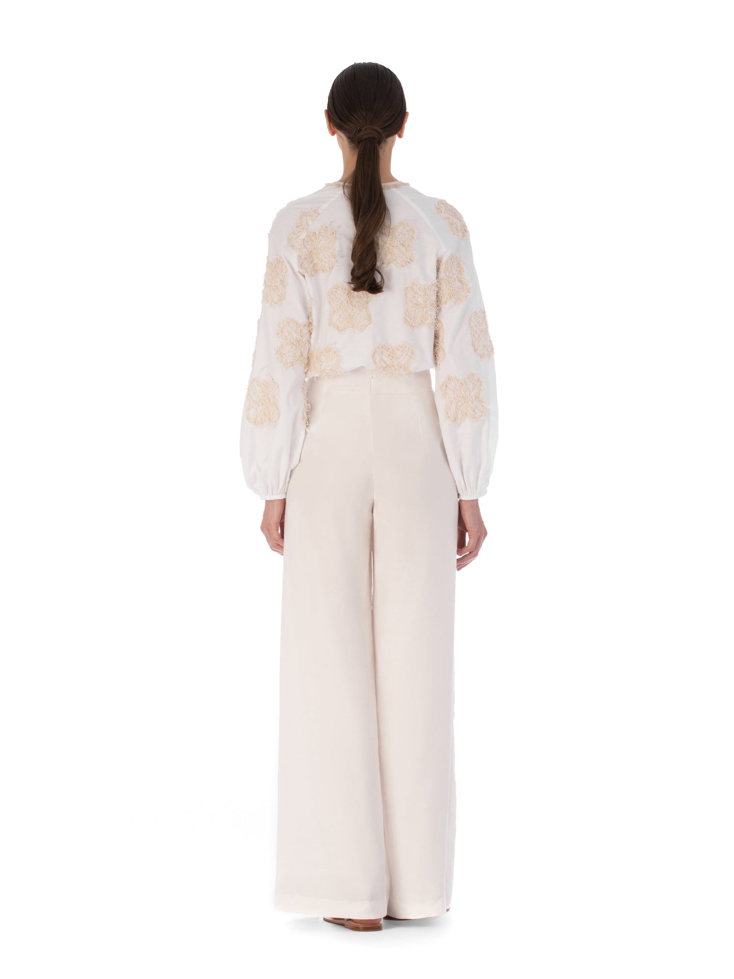 Rear view of a person with long hair wearing the Molveno Blouse Cream Flowers, featuring embroidered floral motifs, paired with high-waisted, wide-leg pants.