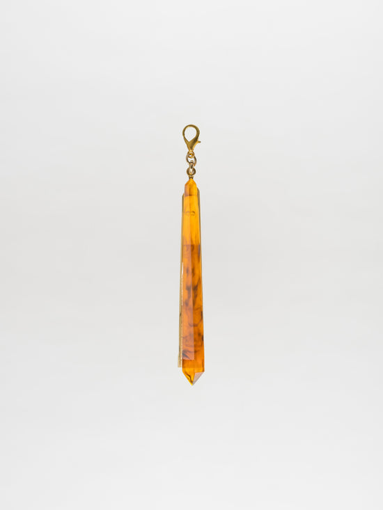 The Montse Charm Amber, an elongated crystal pendant with an amber hue and a metal clasp, is elegantly showcased against a plain white background, highlighting the sophisticated Tcherassi design.