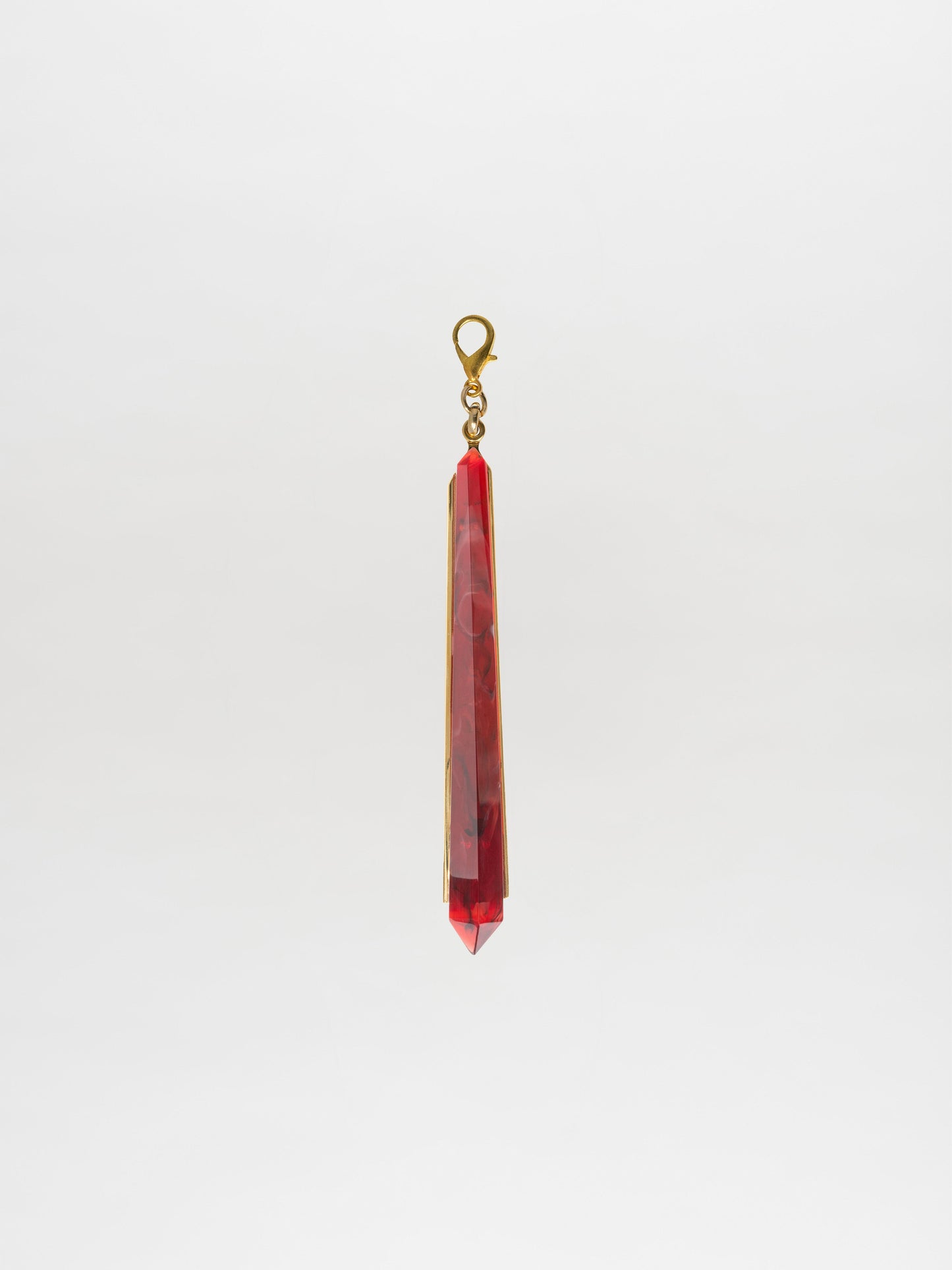 The Montse Charm Rouge, a geometric pendant featuring a red crystal and a gold-plated brass clasp, is elegantly displayed against a plain white background.