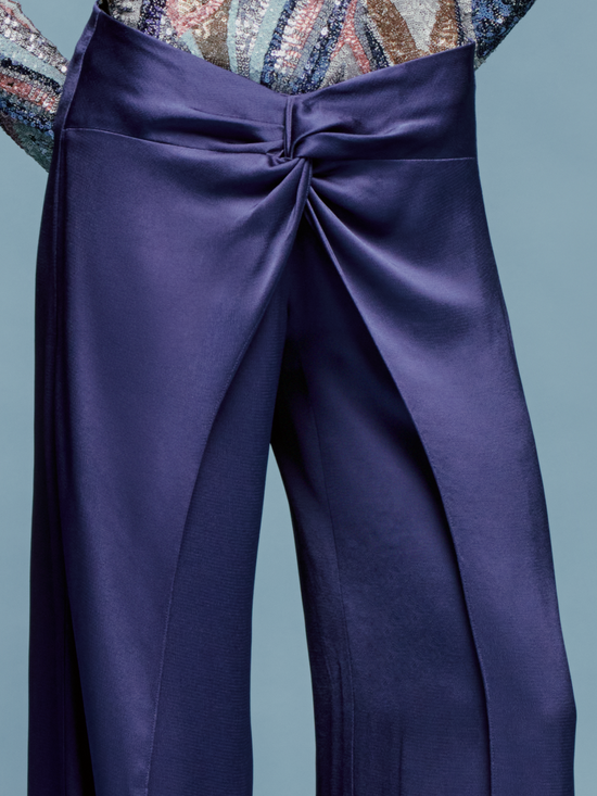 A person stands against a light blue background wearing a patterned long-sleeve top and the high-waisted, wide-leg Nadira Pant in Navy, ready for shipping by August 2024.