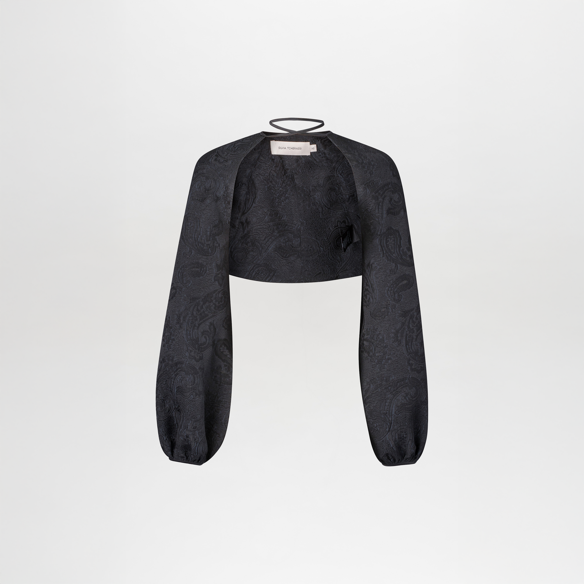 The Nana Cape is a black cropped bolero jacket featuring long, loose sleeves, a subtle jacquard paisley pattern, and a thin neckline strap, shown against a plain white background.