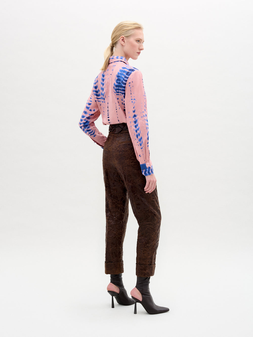 Against a plain backdrop, someone wears the Moad Pant—high-waisted and cropped in Italian fabric with a straight fitted leg—paired with a pink patterned shirt, large gold belt, and black heeled boots.