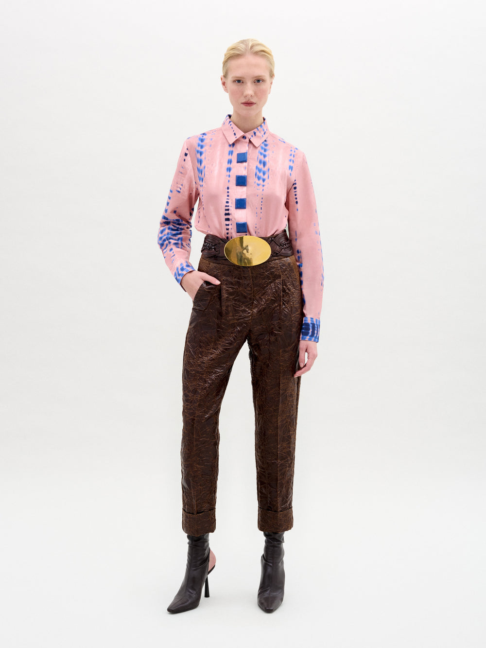 A person stands against a plain background wearing the Moad Pant, a high-waisted cropped style with a straight fitted leg made from Italian fabric, paired with a pink patterned shirt, large gold belt, and black heeled boots.