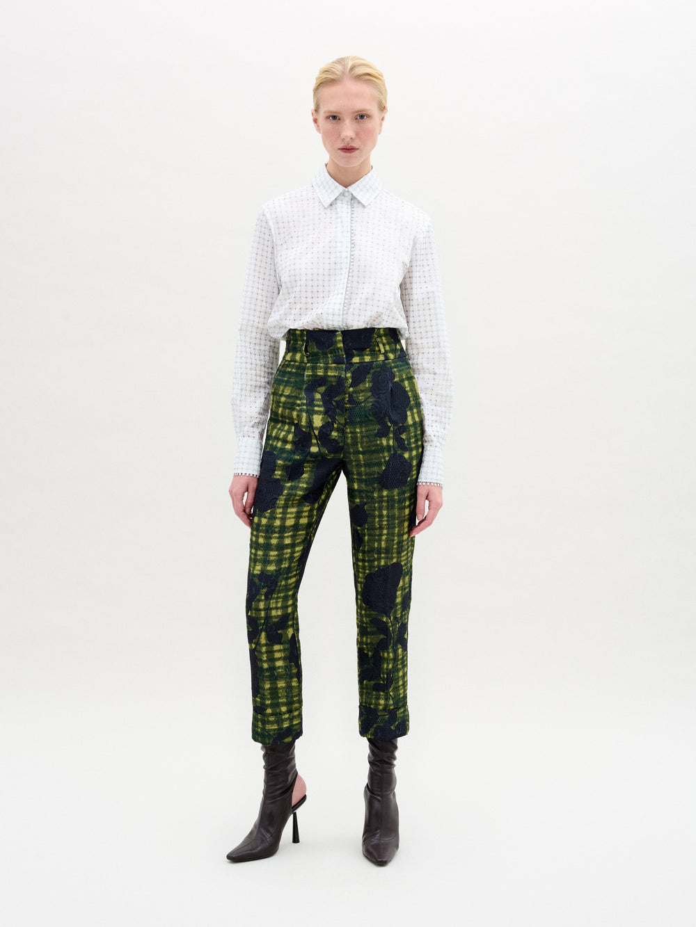 Against a neutral backdrop, a person models a chic outfit with the Naroa Blouse in Italian white patterned fabric, styled with green and blue patterned pants. The ensemble is completed by sleek black high-heeled boots.