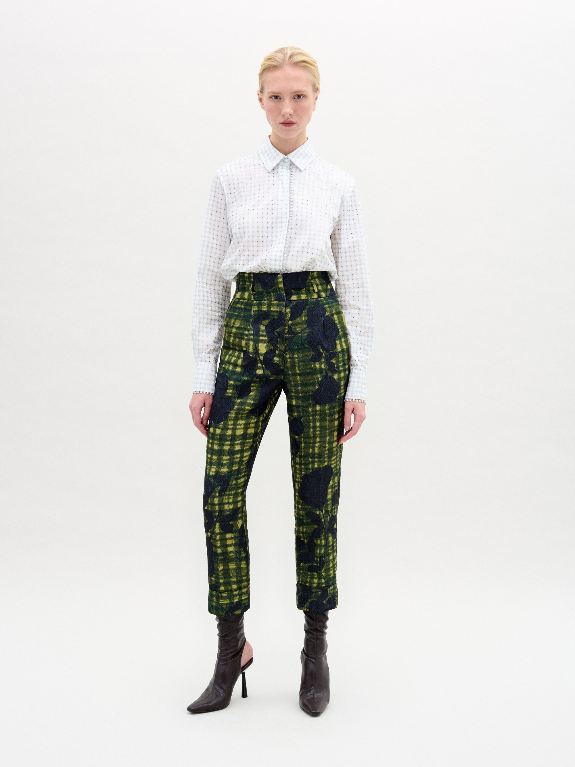 Against a neutral backdrop, a person models a chic outfit with the Naroa Blouse in Italian white patterned fabric, styled with green and blue patterned pants. The ensemble is completed by sleek black high-heeled boots.