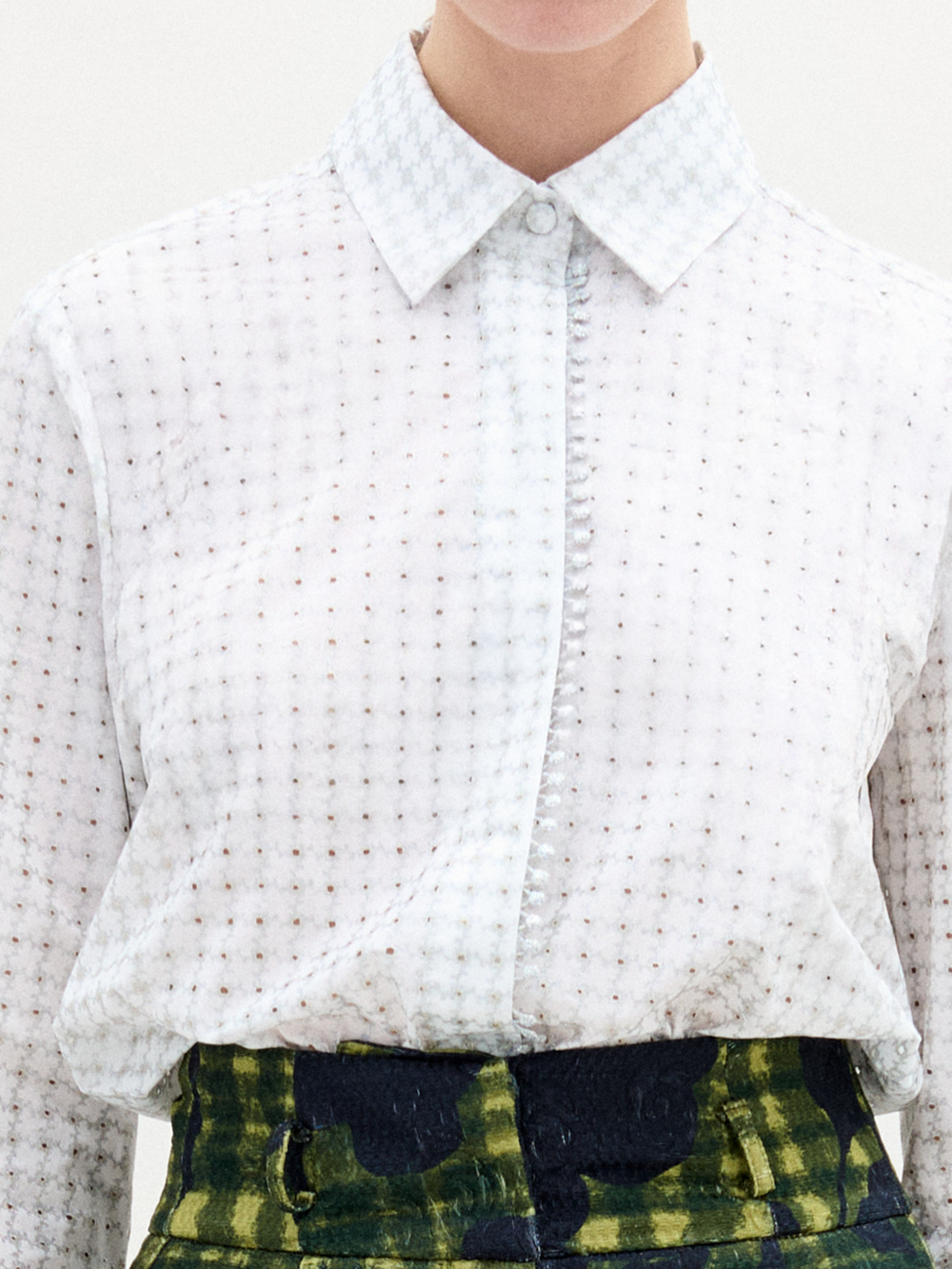 Against a neutral backdrop, a person models a chic outfit with the Naroa Blouse in Italian white patterned fabric, styled with green and blue patterned pants. The ensemble is completed by sleek black high-heeled boots.