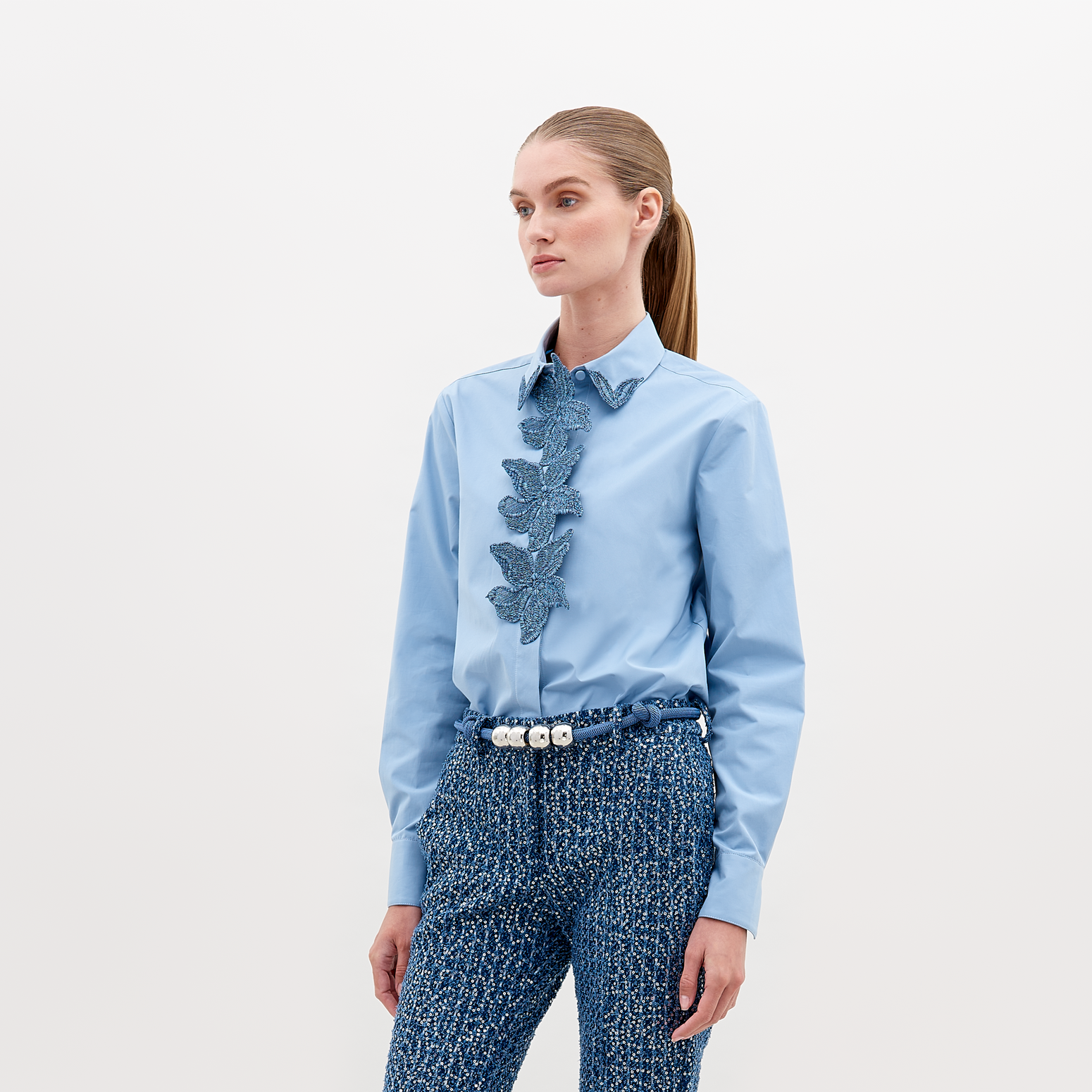 A woman with straight hair in a ponytail wears a Naroa Shirt Celestial Blue with ruffled detailing on the front and textured blue high-waisted pants, accessorized with a white beaded belt. Available for pre-order now, with an expected ship date in November 2024.