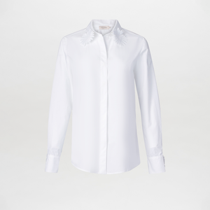 The Naroa Blouse is a white cotton top with long sleeves, a classic collar, and delicate embroidered floral details on the collar tips. Made in Colombia, it’s shown against a plain white background.