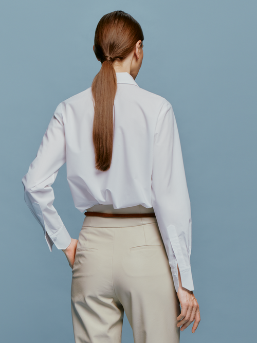 A woman with light brown hair is wearing the Naroa Blouse White, which features a black and gray vertical design, paired with beige pants. She is posing against a blue background in a snapshot taken on September 2nd, 2024.