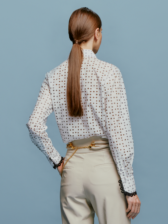 A woman stands against a blue background, wearing the white Naroa Blouse with black dots and light gray pants, accessorized with a chain belt, ready for the fresh styles of September 2nd, 2024.