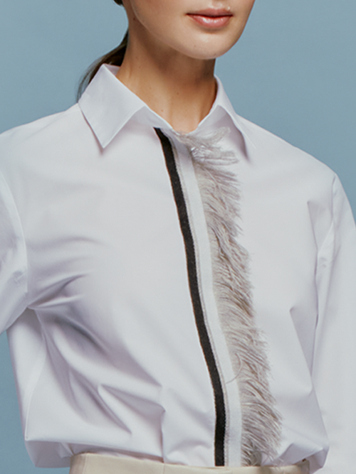 A woman with light brown hair is wearing the Naroa Blouse White, which features a black and gray vertical design, paired with beige pants. She is posing against a blue background in a snapshot taken on September 2nd, 2024.