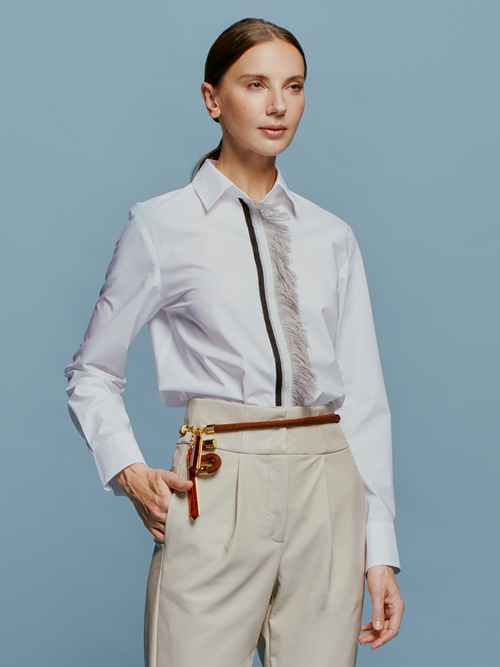 A woman with light brown hair is wearing the Naroa Blouse White, which features a black and gray vertical design, paired with beige pants. She is posing against a blue background in a snapshot taken on September 2nd, 2024.