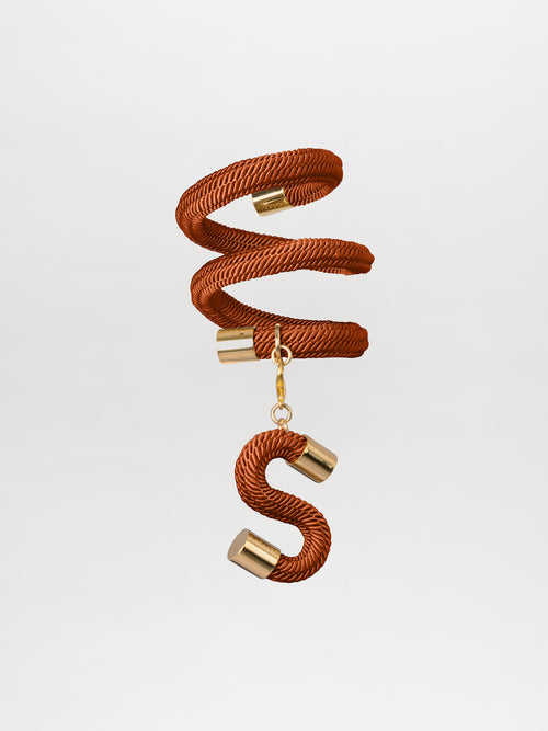 The Nicky Bracelet Ochre, featuring a spiral rope design with gold-plated brass accents and an "S" charm in a Tcherassi style, hangs suspended in mid-air against a plain background.