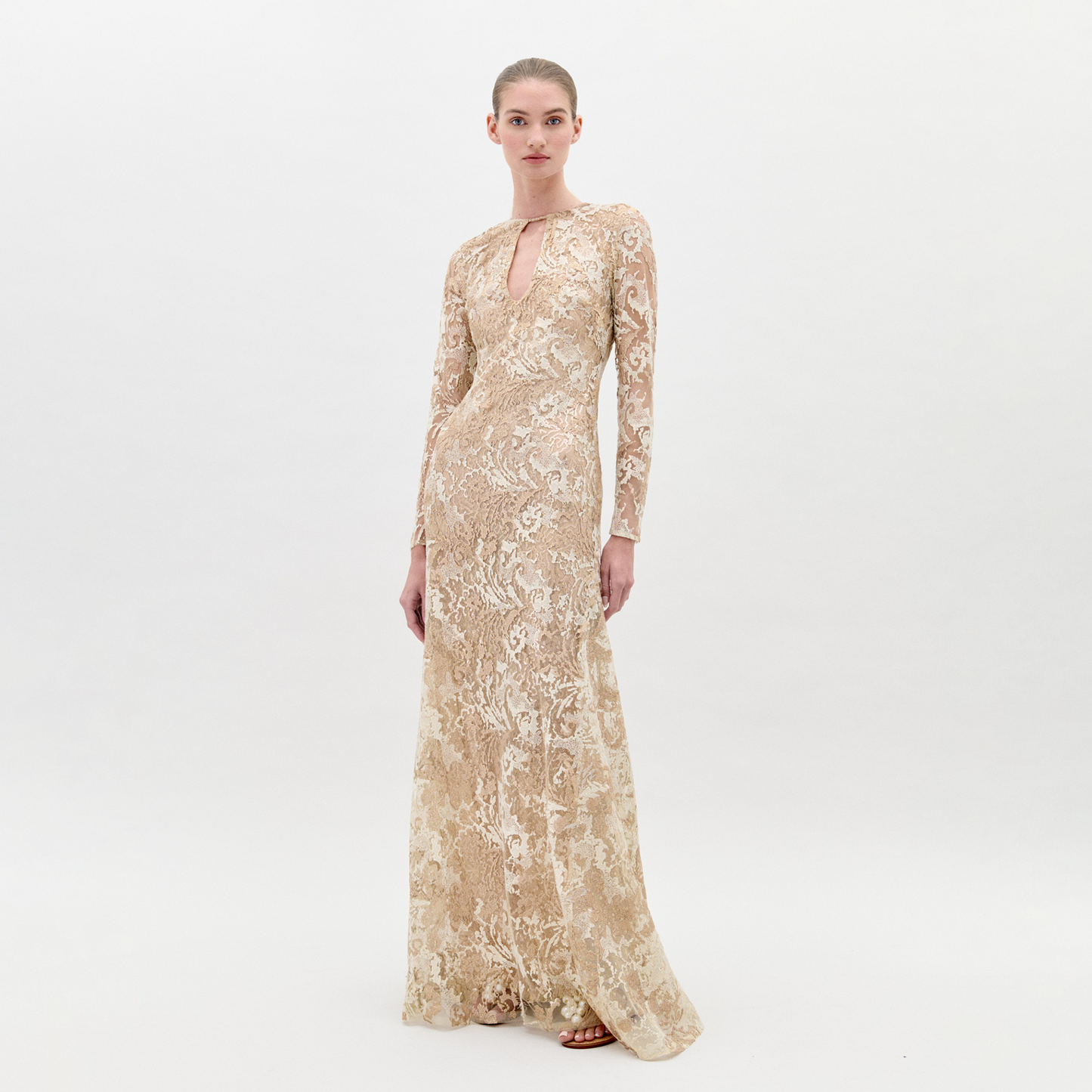 A woman in the Nila Dress Gold, a beige, floor-length, long-sleeved lace gown with a keyhole neckline, stands against a plain background, ready for pre-order ahead of the ship-by date on February 1st, 2025.
