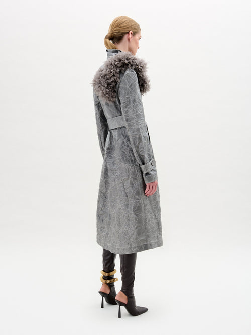 The model wears the Nilo Trench, a textured gray trench coat made from Italian fabric with a floral embellishment on the shoulder, cinched by a gray belt. It is styled with black pants and high-heeled boots against a plain white background.