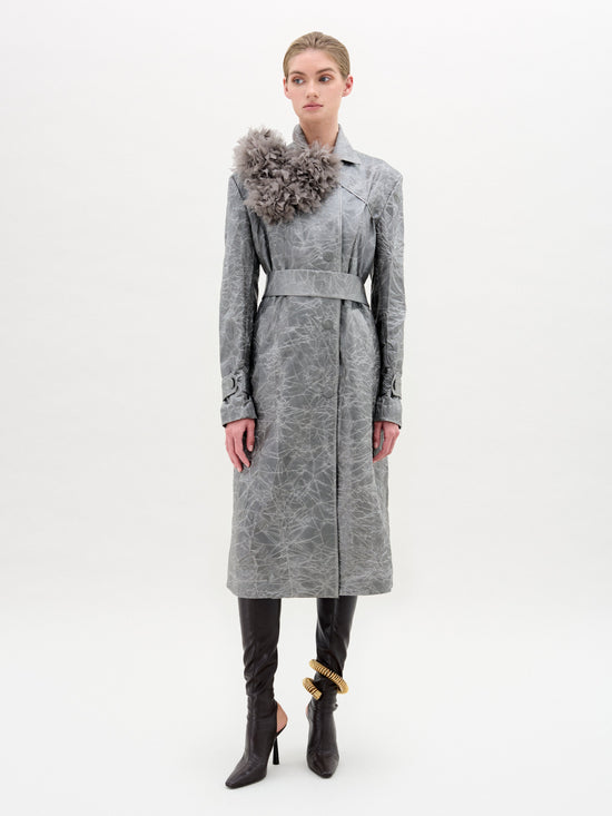 The model wears the Nilo Trench, a textured gray trench coat made from Italian fabric with a floral embellishment on the shoulder, cinched by a gray belt. It is styled with black pants and high-heeled boots against a plain white background.