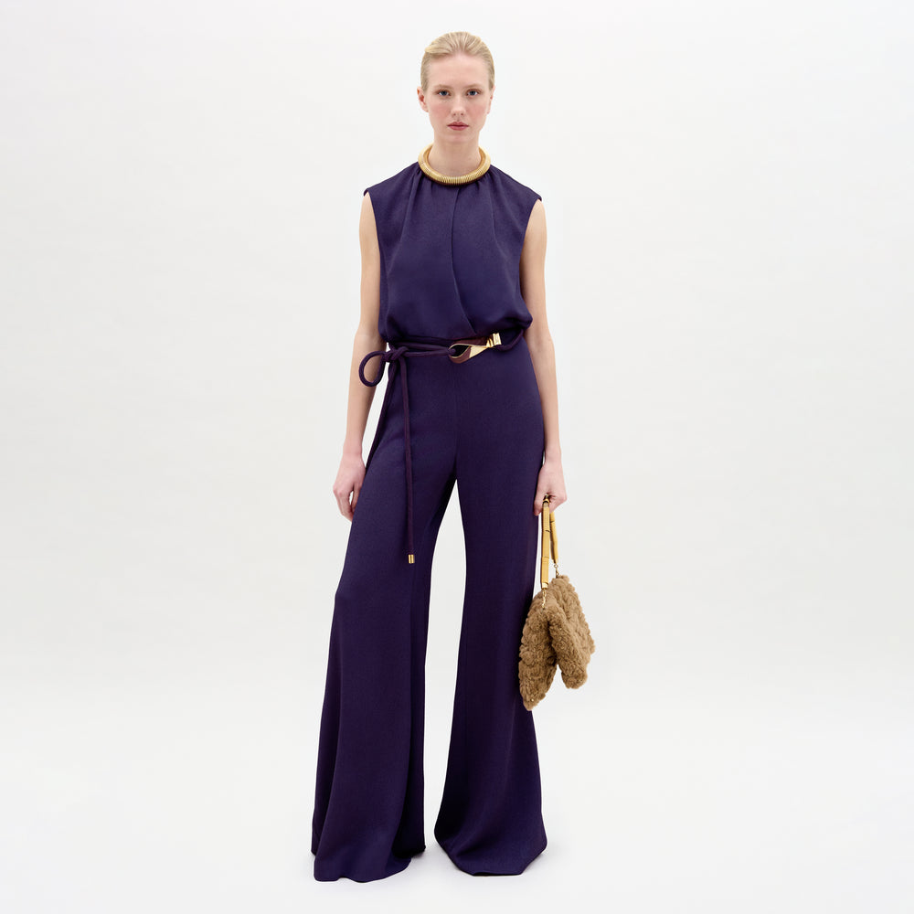 A woman wearing the Noel Jumpsuit in color Purple with a Turni clutch in color Camel on a white background.
