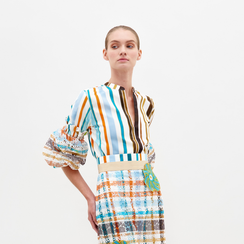 A model, dressed in the Nuru Blouse featuring multi-colored stripes and puffy sleeves, along with a patterned skirt, poses against a plain background to highlight fashion available for pre-order.