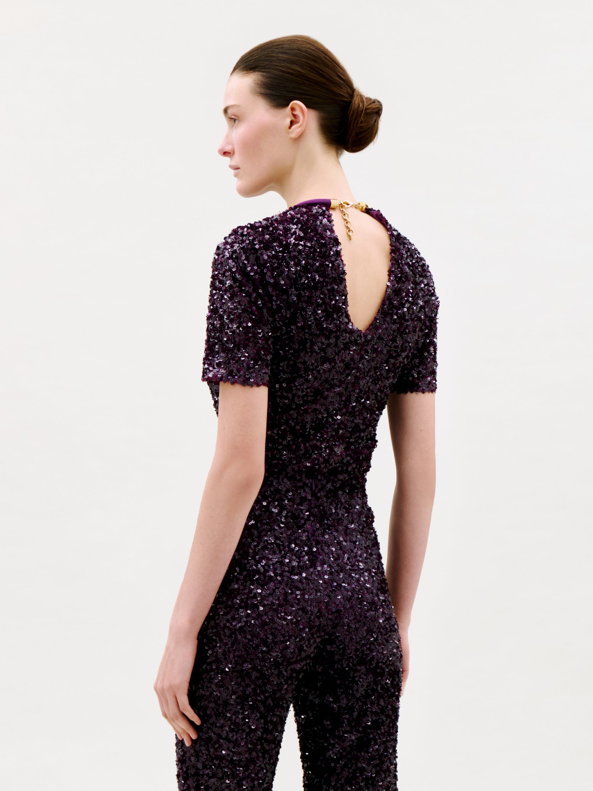 A person with brunette hair in a low bun models the Napoles Blouse, a dark purple sequin top featuring a fitted silhouette, short sleeves, and a keyhole opening in the back, standing against a plain light background.