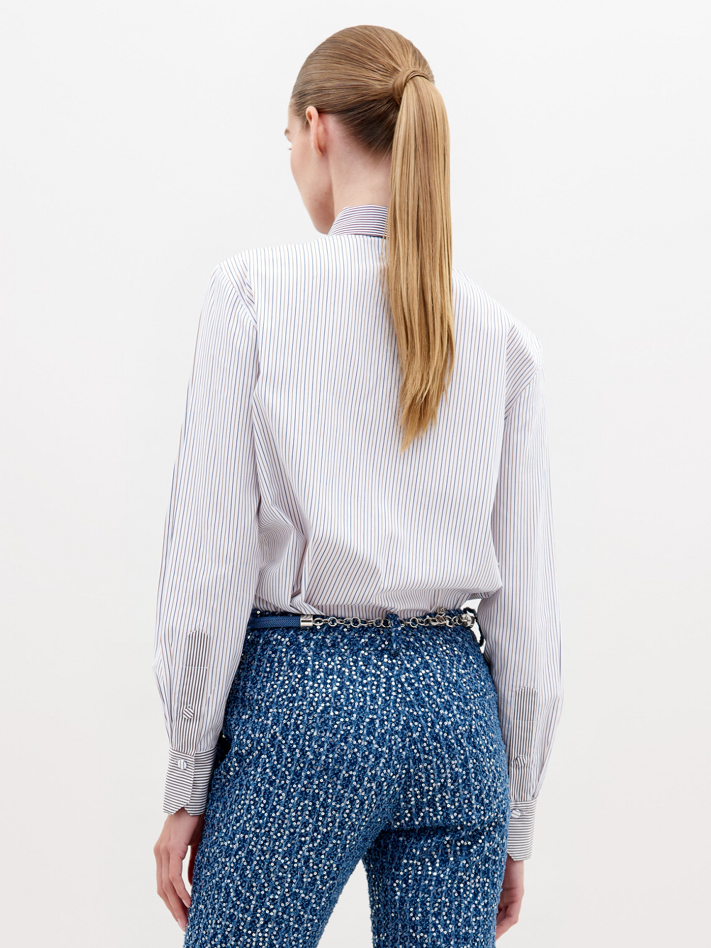 A person with blonde hair slicked back wears a white shirt with vertical stripes and a detailed blue lace front, along with textured blue pants and a belt with large pearl-like embellishments. Now available for pre-order, the Naroa Shirt Blue Brown Stripes will ship by 2024.