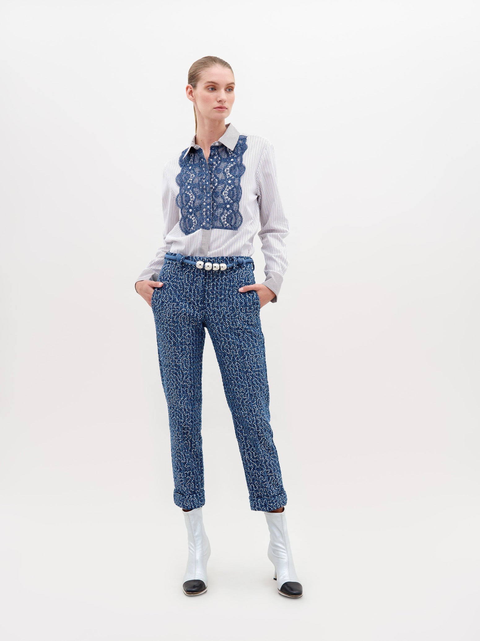 A woman stands against a plain background wearing a white blouse with blue lace detail, Garmet Pant Navy Sequins, a pearl belt, and white ankle boots with black tips. This stylish ensemble is available for pre-order now and will ship by November 15th, 2024.