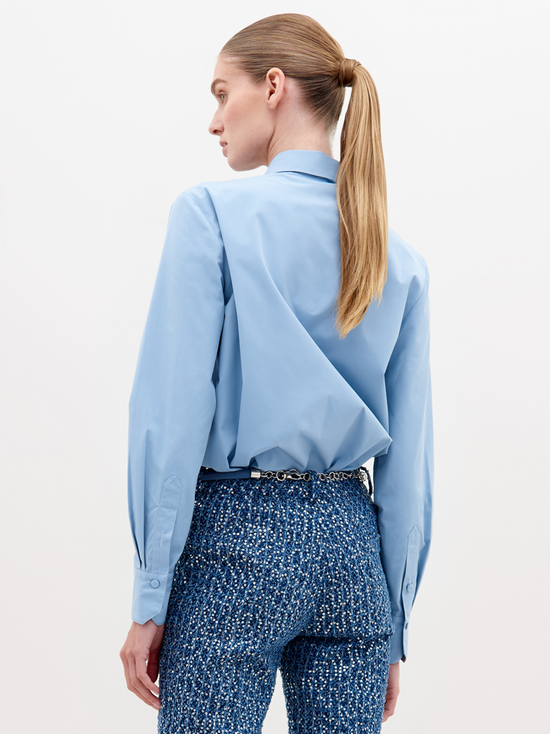 A woman with straight hair in a ponytail wears a Naroa Shirt Celestial Blue with ruffled detailing on the front and textured blue high-waisted pants, accessorized with a white beaded belt. Available for pre-order now, with an expected ship date in November 2024.