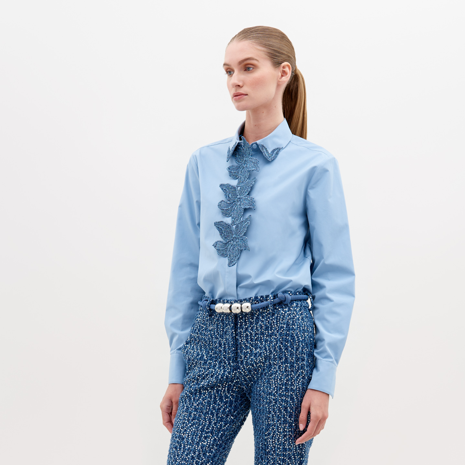 A woman with straight hair in a ponytail wears a Naroa Shirt Celestial Blue with ruffled detailing on the front and textured blue high-waisted pants, accessorized with a white beaded belt. Available for pre-order now, with an expected ship date in November 2024.