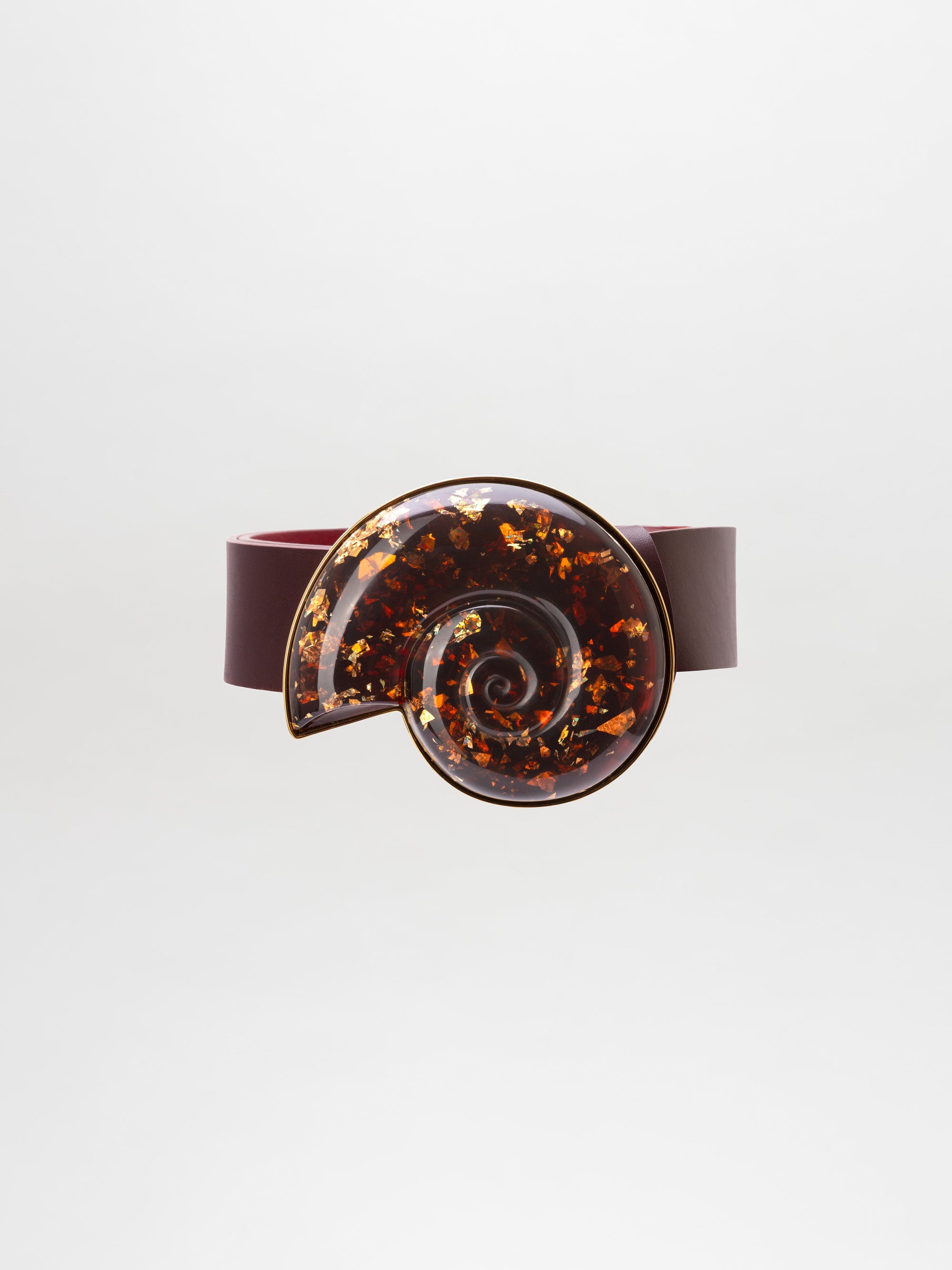 The Nautilus Belt is a dark maroon accessory with a large, glossy, spiral-shaped buckle adorned with amber and gold flecks.