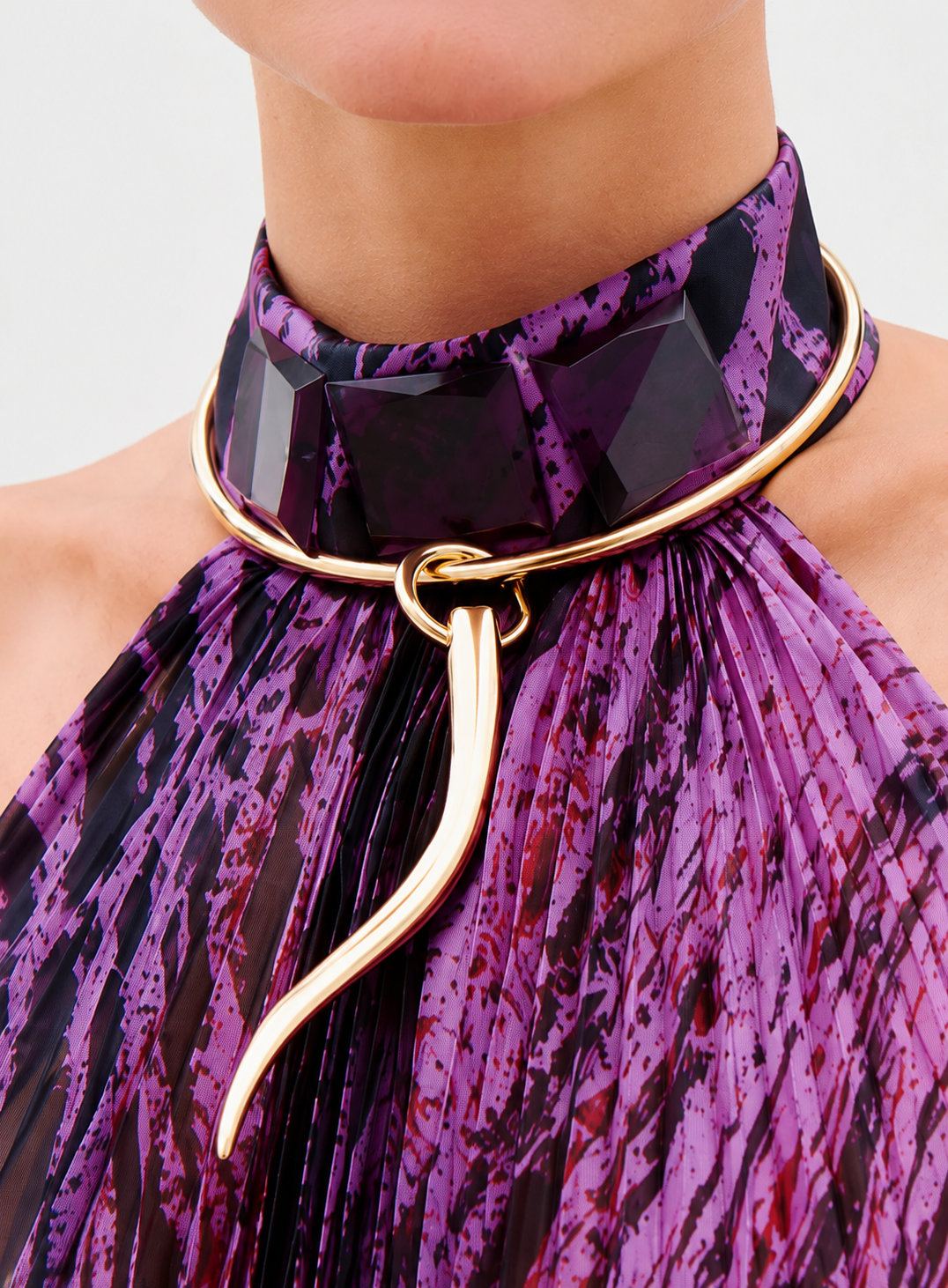 A person wearing the Melinda Choker, featuring faceted purple accents, along with a gold-plated sculptural necklace with an adjustable length over a pleated top with a purple and black pattern.