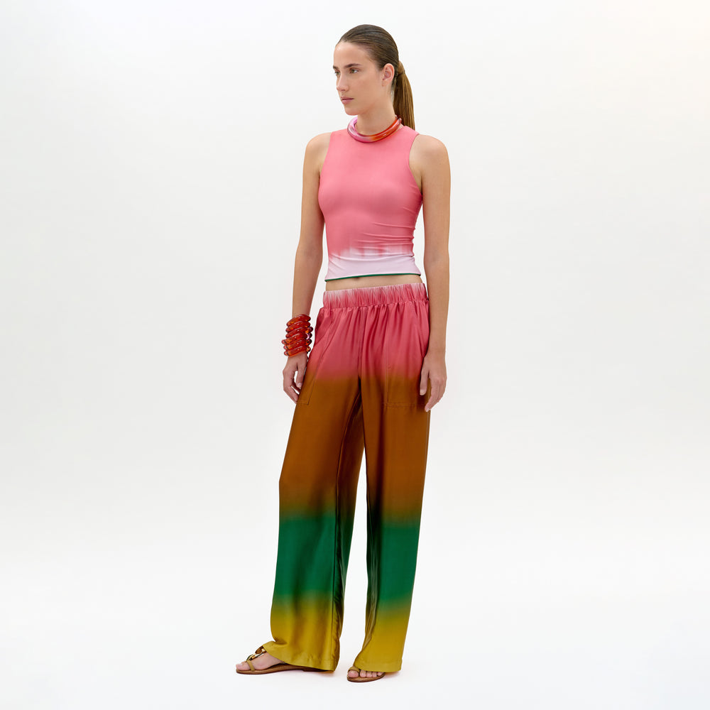 A woman stands against a plain background, wearing the Nerea Pant—loose, high-waisted with an elastic waistband in a pink, brown, green, and yellow Italian fabric—paired with a sleeveless pink top, red bracelet, and flat sandals.