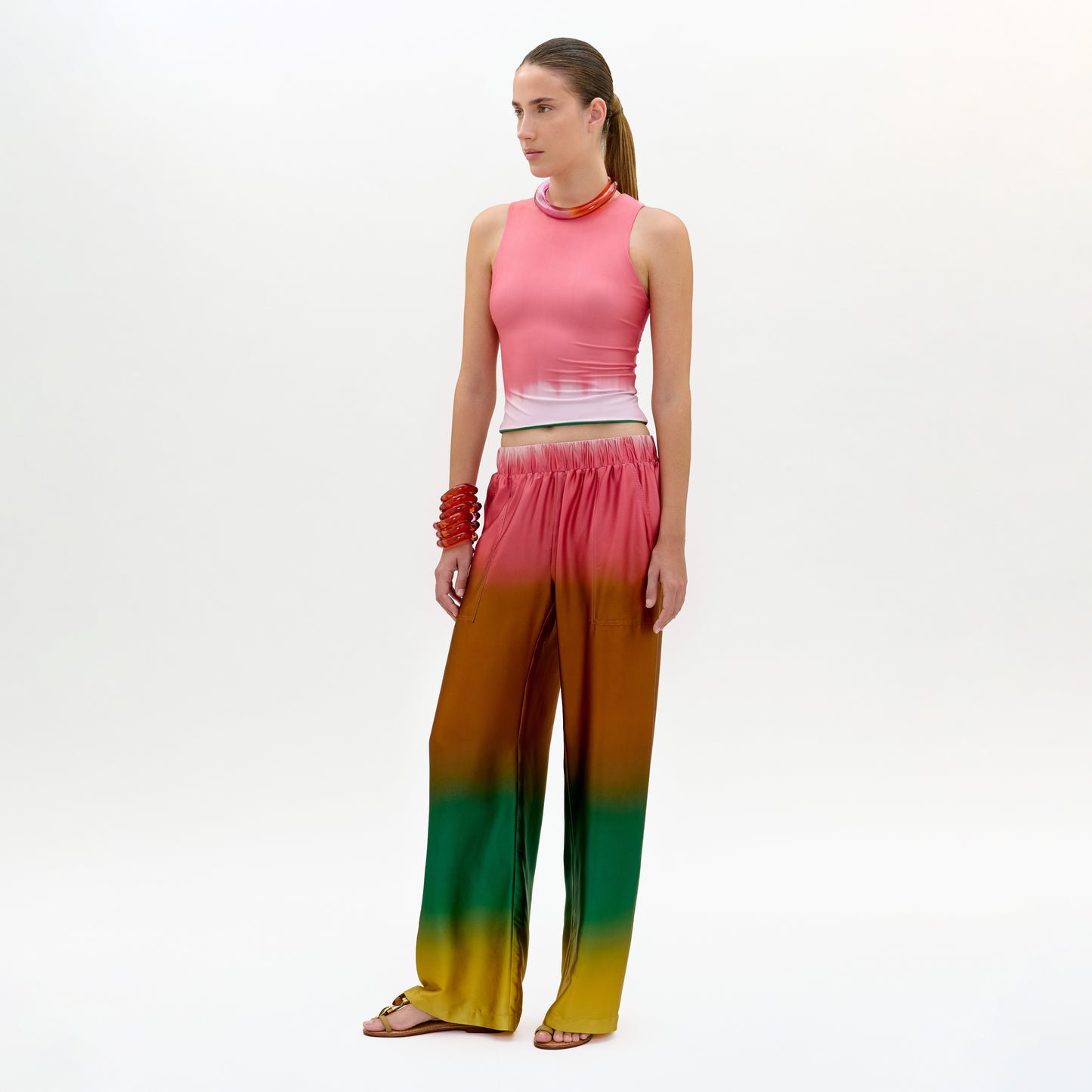 A woman stands against a plain background, wearing the Nerea Pant—loose, high-waisted with an elastic waistband in a pink, brown, green, and yellow Italian fabric—paired with a sleeveless pink top, red bracelet, and flat sandals.