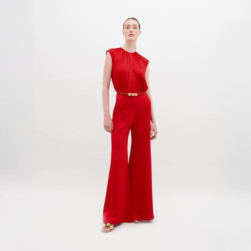 A person stands against a plain background, wearing a Noel Jumpsuit Rouge with a gold-buckle belt and open-toed shoes. This stylish ensemble will be available for pre-order and is expected to ship by November 15th, 2024.