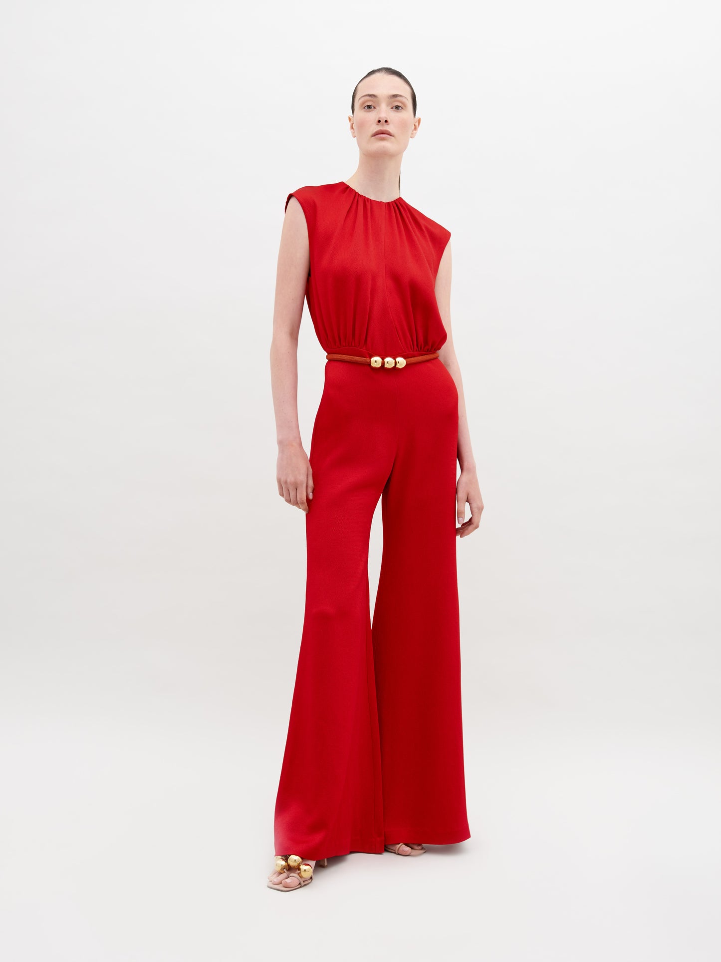 A person stands against a plain background, wearing a Noel Jumpsuit Rouge with a gold-buckle belt and open-toed shoes. This stylish ensemble will be available for pre-order and is expected to ship by November 15th, 2024.