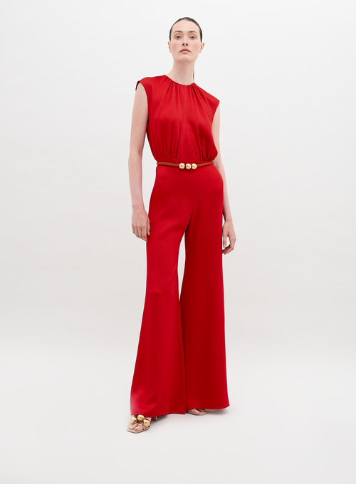 A person stands against a plain background, wearing a Noel Jumpsuit Rouge with a gold-buckle belt and open-toed shoes. This stylish ensemble will be available for pre-order and is expected to ship by November 15th, 2024.