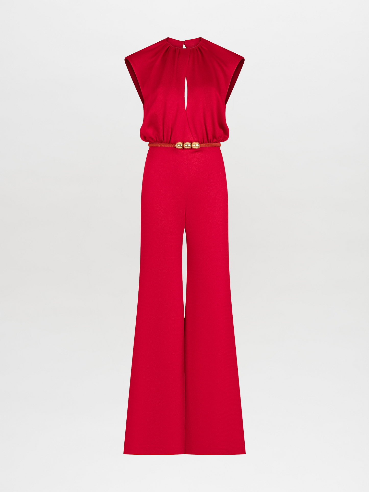 A person stands against a plain background, wearing a Noel Jumpsuit Rouge with a gold-buckle belt and open-toed shoes. This stylish ensemble will be available for pre-order and is expected to ship by November 15th, 2024.