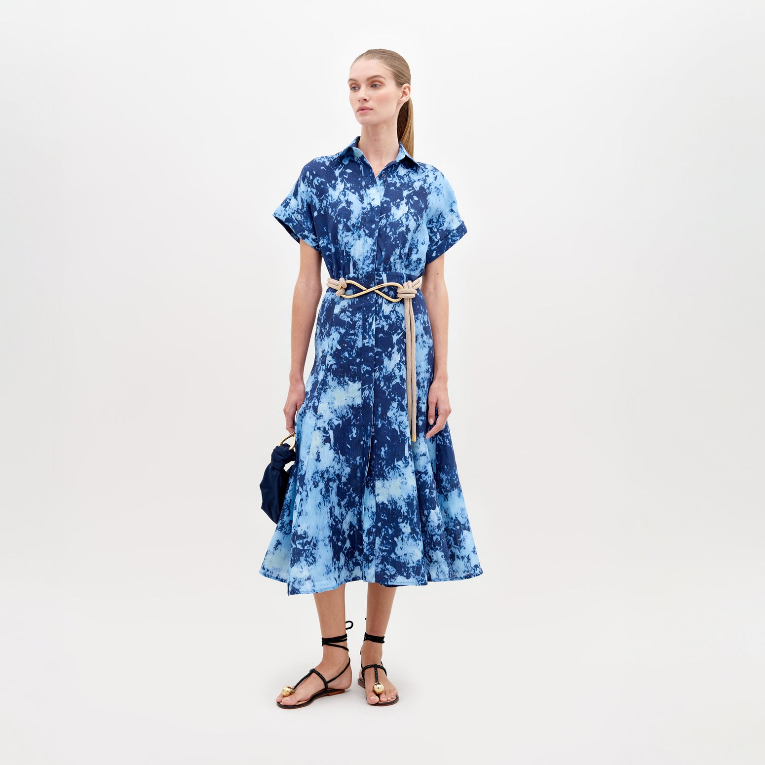 A woman stands against a white background, wearing a Noor Dress Indigo with a beige belt and black sandals, holding a dark blue bag—a stylish ensemble available for pre-order until November 15th, 2024.