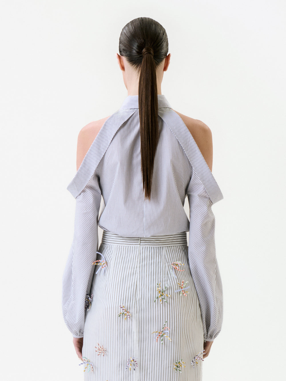 Back view of a person wearing a Nurith Shirt featuring light blue and white stripes with cold-shoulder cutouts and a matching striped skirt with small embroidered floral details.