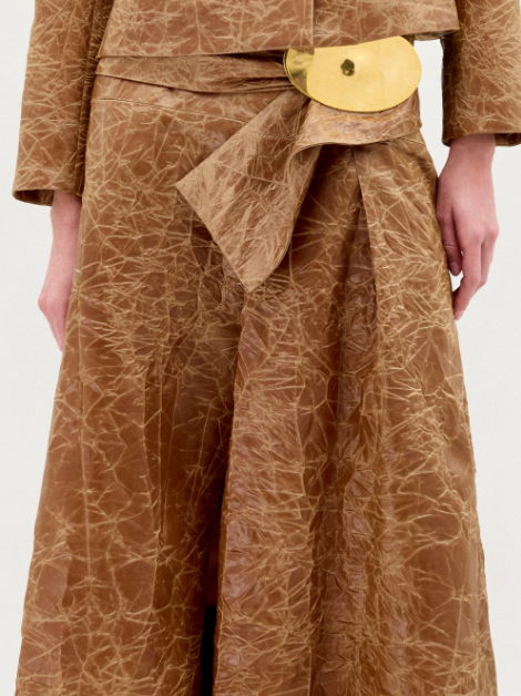 A woman wears the Olga Skirt in brown with a matching textured A-line jacket, gold accent belt, dark ankle boots, and a minimal background.