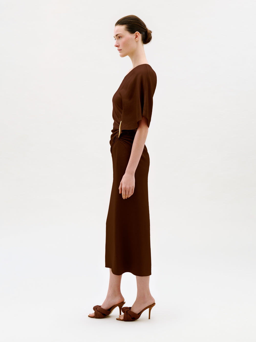 A woman stands in profile against a plain light background, wearing the Oby Dress—a brown short-sleeved midi dress crafted from luxurious Italian fabric—paired with brown heeled sandals. Her hair is tied back in a neat bun.
