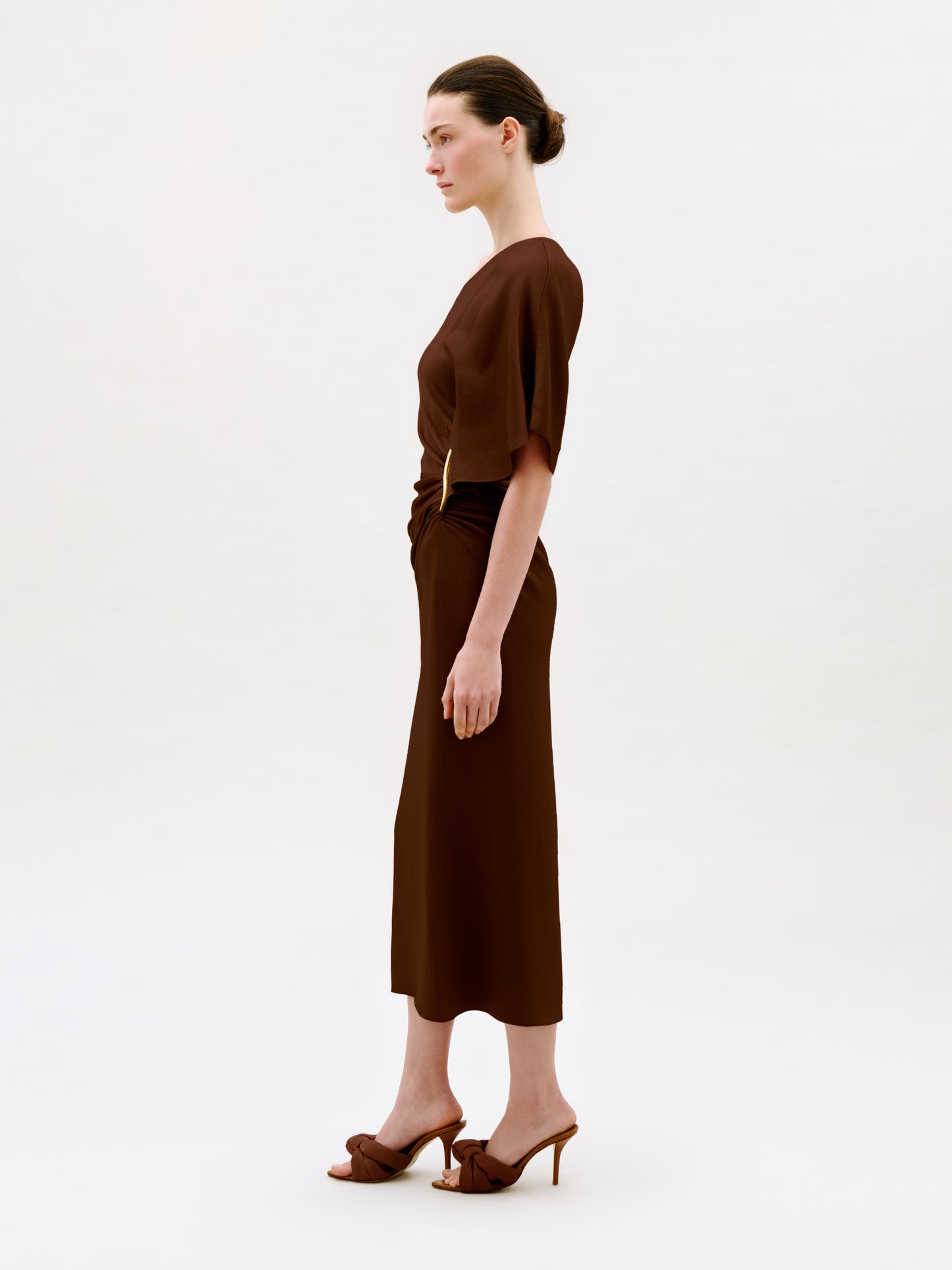 A woman stands in profile against a plain light background, wearing the Oby Dress—a brown short-sleeved midi dress crafted from luxurious Italian fabric—paired with brown heeled sandals. Her hair is tied back in a neat bun.