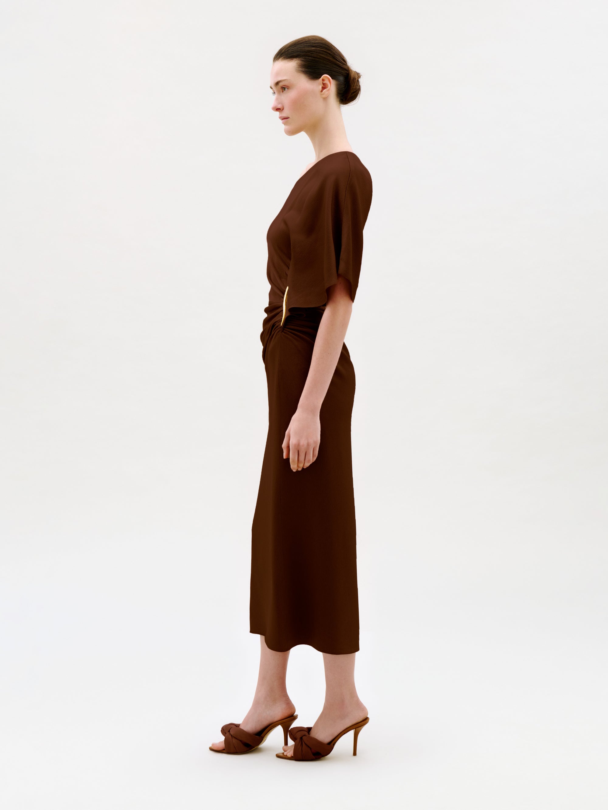 A woman stands in profile against a plain light background, wearing the Oby Dress—a brown short-sleeved midi dress crafted from luxurious Italian fabric—paired with brown heeled sandals. Her hair is tied back in a neat bun.