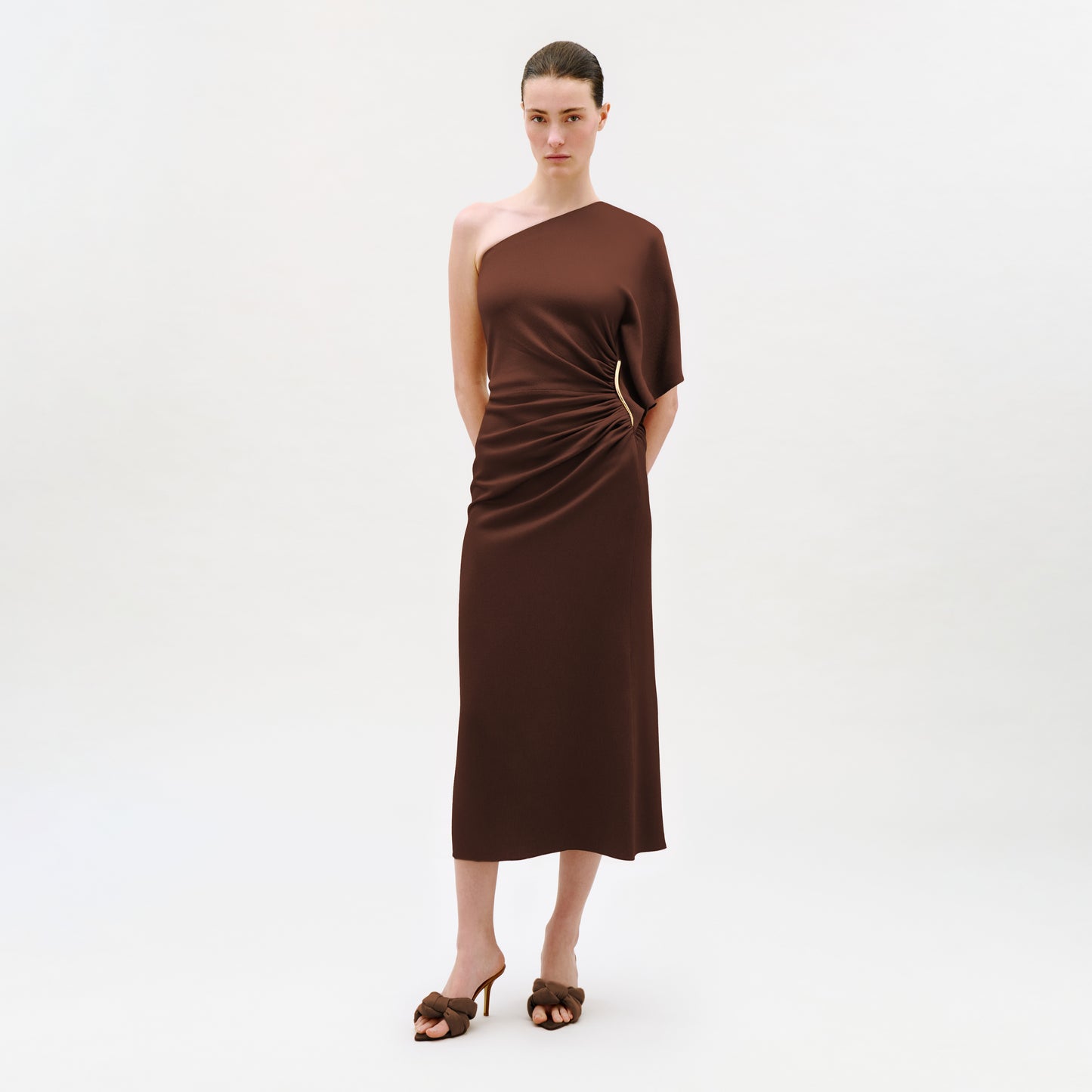 A woman stands against a white background, wearing the Oby Dress—an elegant midi dress with an asymmetric neckline, crafted from luxurious Italian fabric. The brown, one-shoulder design features side ruching and is paired with matching brown heeled sandals. Her hair is pulled back and her hands are behind her back.