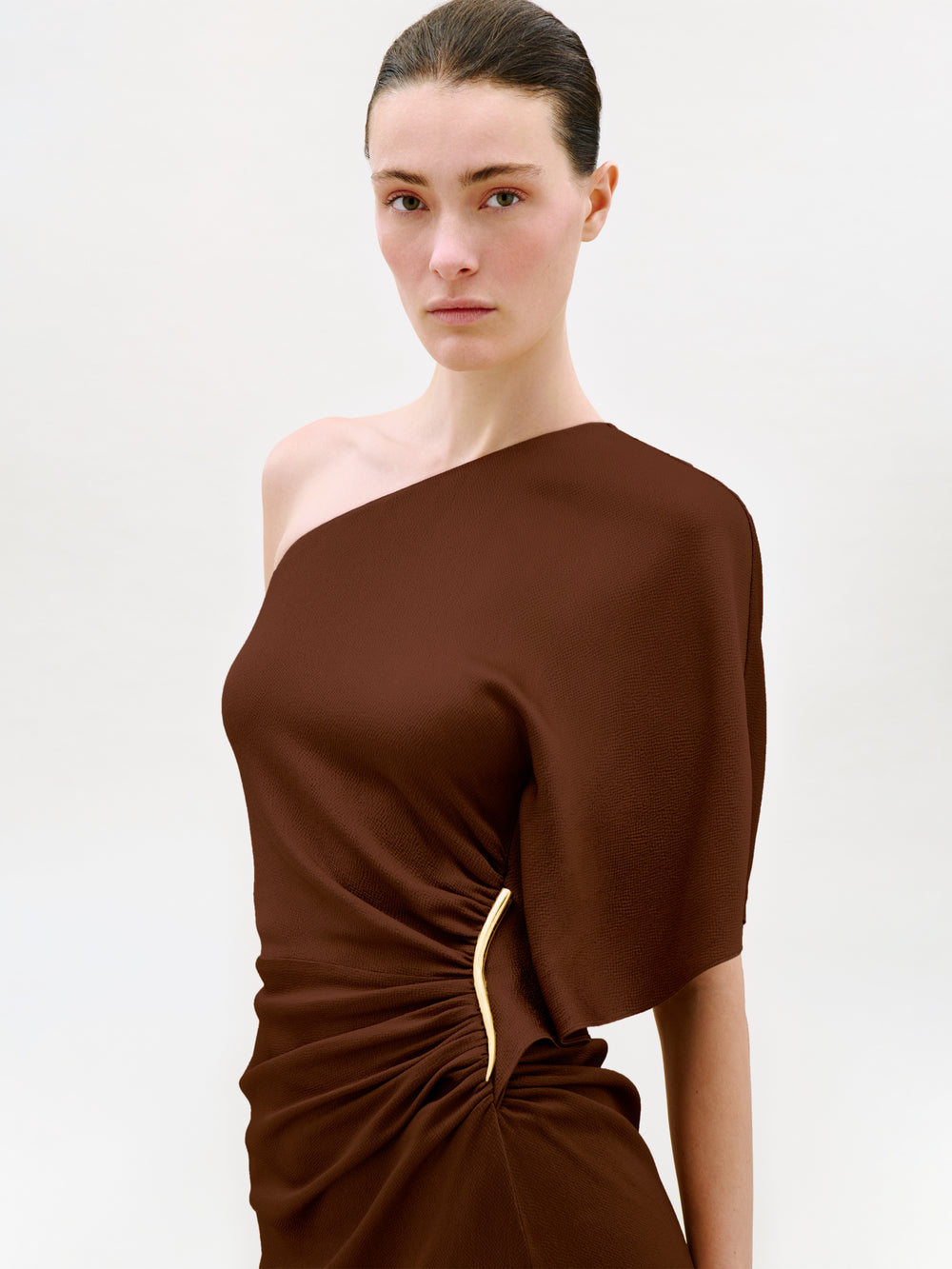 A woman with dark hair in a sleek bun wears the Oby Dress, a brown one-shoulder midi dress crafted from luxurious Italian fabric with a gold accent at the waist, standing against a plain light background.
