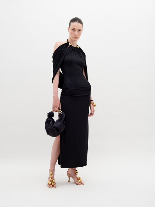 A woman stands against a white background, showcasing the Odele Dress Black with gold accents. She holds a small black bag and wears gold-embellished high heels. Pre-order now to ensure delivery by November 15th, 2024.